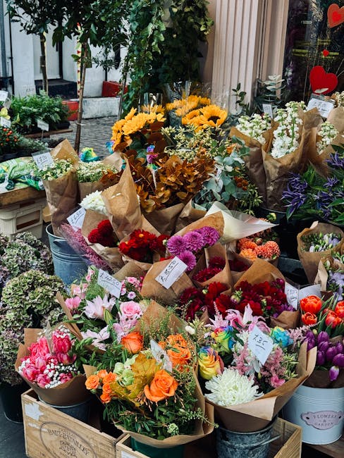 A vibrant display of fresh flowers arranged in paper wrapping and placed on a wooden stand outside a flower shop. The arrangement includes a variety of blooms such as bright yellow sunflowers, pink li