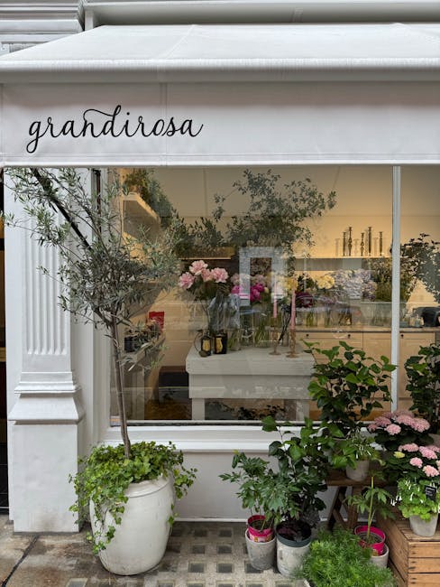 A storefront window of 'grandirose' with a white awning, displaying an arrangement of fresh pink peonies and other floral elements inside. Outside, in front of the window, there are potted greenery in