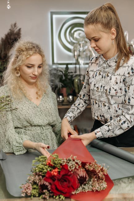 Two women working together on a floral arrangement in a well-lit shop environment. The older woman, with curly blonde hair, is wearing a light green dress with a subtle pattern, while the younger woma