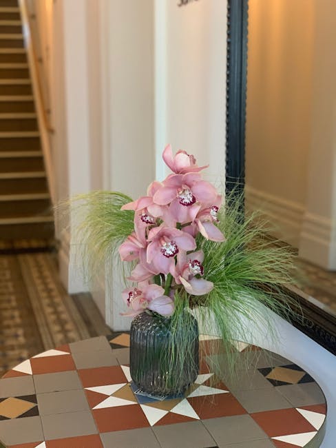 A tall floral arrangement featuring soft pink orchids with multiple blooms on each stem, arranged in a clear, fluted glass vase. The orchids are complemented by wispy green foliage and delicate grass-