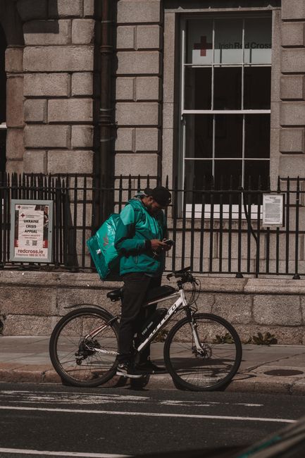 The image shows a person holding a smartphone, standing next to a bicycle on a city sidewalk in front of a stone building with a large window. The individual is wearing a teal jacket, holding a large 