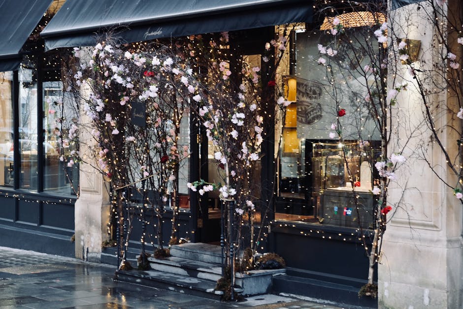 The storefront of Marylebone shopfront floral displays decorated with intertwined bare branches draped in numerous white blossoms and accented with a few red flowers, illuminated by warm string lights