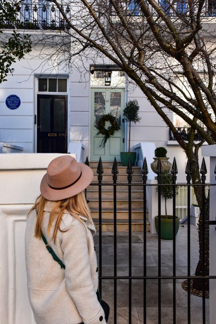 A woman wearing a beige coat and a wide-brimmed pink hat stands in front of a black wrought iron gate, facing a white townhouse entrance decorated with a green wreath on the door. The entrance feature