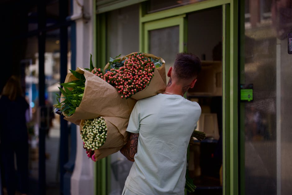 A person with short dark hair and a white t-shirt is holding a large bouquet of fresh flowers wrapped in brown paper, positioned over their shoulder in front of a green storefront window. The bouquet