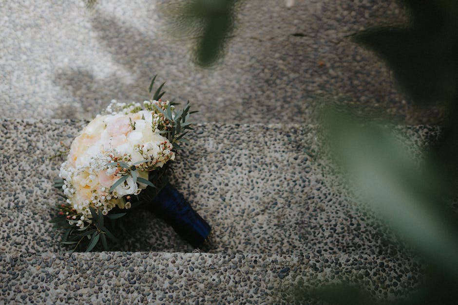 A close-up of a bridal bouquet of fresh flowers, including white and pale pink roses, white lisianthus, and small white filler flowers, accented with sprigs of greenery such as eucalyptus leaves. The