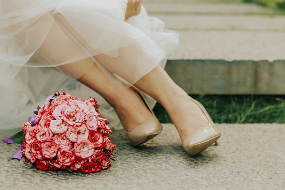 Close-up of a bride's legs and feet wearing beige high-heeled shoes, seated outdoors on a textured rug. Next to her, a large bouquet of pink roses and smaller purple flowers is resting on the ground,